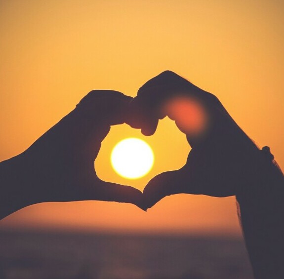 Hands in the shape of a heart with the sun in the background centered in the middle. 